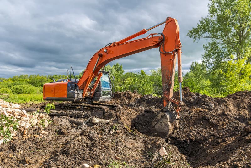 Large orange crawler excavator is excavating digging a trench against backdrop of green trees and an overcast dramatic summer sky. Large orange crawler excavator is excavating digging a trench against backdrop of green trees and an overcast dramatic summer sky.