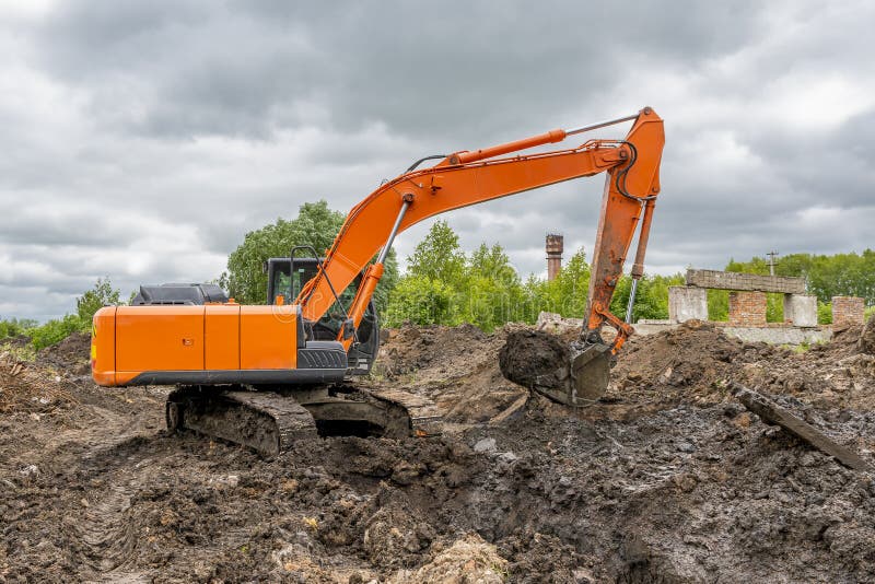 Large orange crawler excavator is excavating digging a trench against backdrop of green trees and an overcast dramatic summer sky. Large orange crawler excavator is excavating digging a trench against backdrop of green trees and an overcast dramatic summer sky.