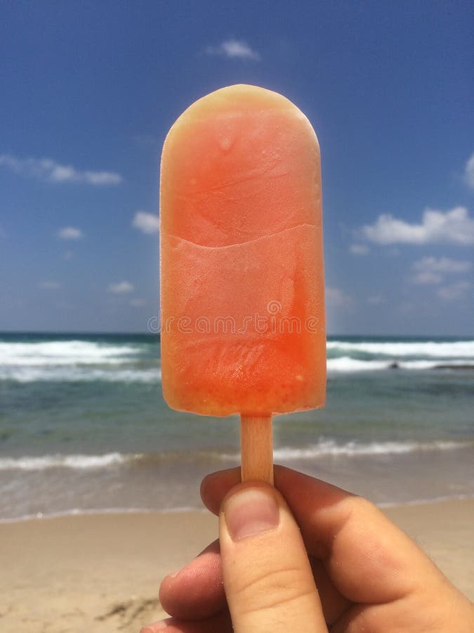 Hand holding an orange popsicle with the ocean in the background. Hand holding an orange popsicle with the ocean in the background