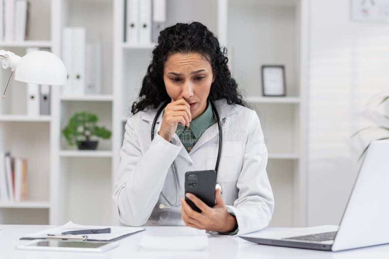 A Hispanic woman, dressed as a doctor, appears anxious while looking at a text message on her smartphone in a medical office setting. A Hispanic woman, dressed as a doctor, appears anxious while looking at a text message on her smartphone in a medical office setting.