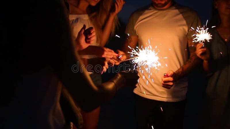 Os amigos que estão em torno de um fogo mais claro durante a noite party no beira-mar para iluminar luzes dos chuveirinhos de ben