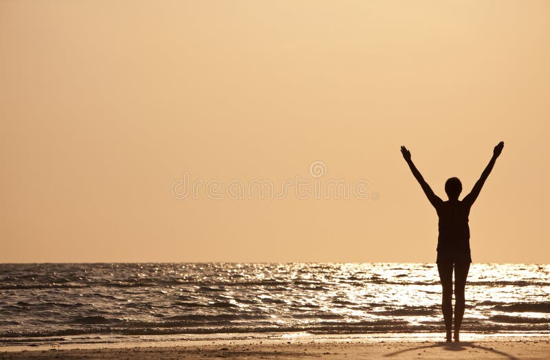 A young woman standing arms raised at sunset or sunrise on a beach. A young woman standing arms raised at sunset or sunrise on a beach