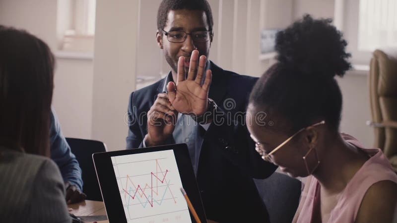 Os executivos afro-americanos falam, mostram gestos e comunicam-se na reunião do escritório, olhando diagramas do portátil