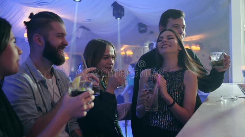 Os homens e as jovens mulheres com cocktail coloridos fazem um brinde e têm o bom tempo perto do contador da barra no clube notur