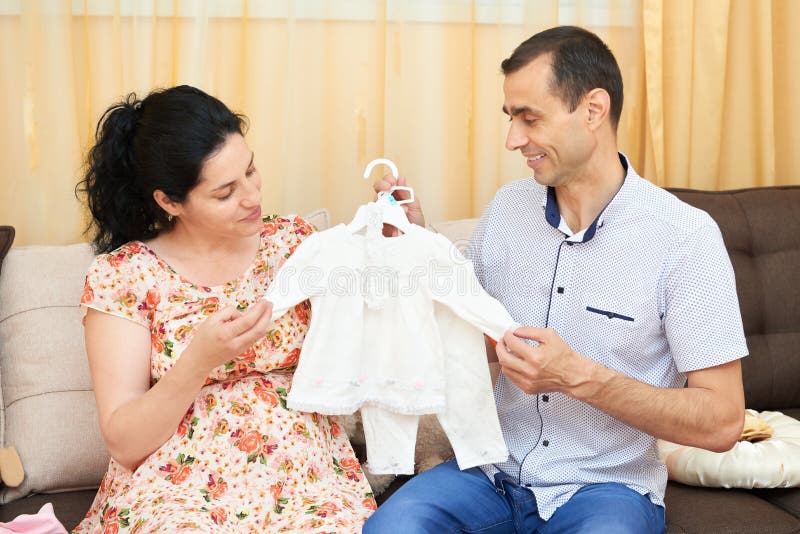 Os pais escolhem a roupa para o bebê recém-nascido Mulher gravida e homem Pares felizes que sentam-se no sofá em casa foto de stock royalty free Os pais escolhem a roupa para o bebê recém-nascido Mulher gravida e homem Pares felizes que sentam-se no sofá em casa foto de stock royalty free
