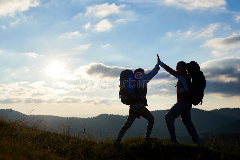 Os pares alegres com as trouxas sobre a montanha dão-se a elevação cinco contra o por do sol imagens de stock Os pares alegres com as trouxas sobre a montanha dão-se a elevação cinco contra o por do sol imagens de stock