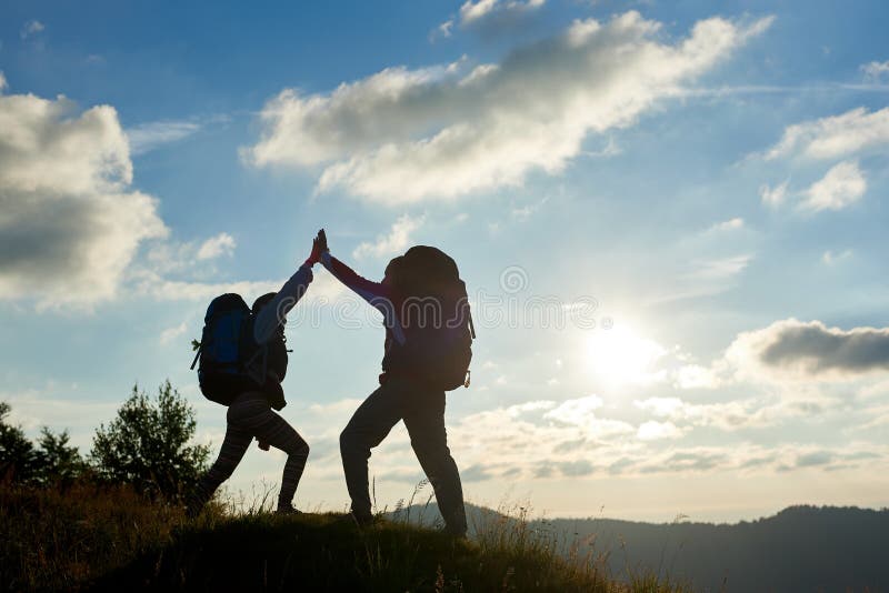 Os pares alegres com as trouxas sobre a montanha dão-se a elevação cinco contra o por do sol imagem de stock Os pares alegres com as trouxas sobre a montanha dão-se a elevação cinco contra o por do sol imagem de stock
