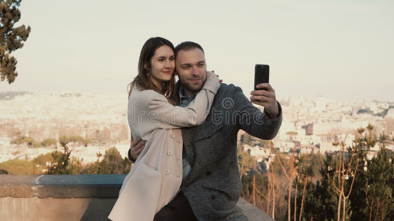 Os pares bonitos novos tomam a foto do selfie contra o panorama de Roma, Itália O homem feliz beija a mulher e os abraços imagens de stock Os pares bonitos novos tomam a foto do selfie contra o panorama de Roma, Itália O homem feliz beija a mulher e os abraços imagens de stock