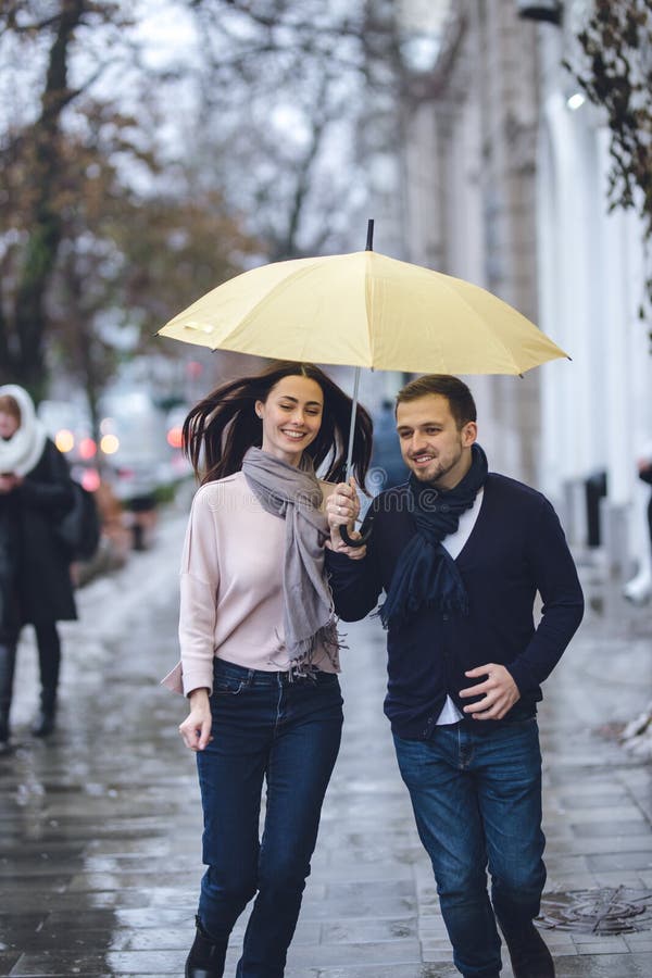 Os pares bonitos, o indivíduo e sua amiga vestidos na roupa ocasional estão correndo sob o guarda-chuva na rua no imagem de stock royalty free Os pares bonitos, o indivíduo e sua amiga vestidos na roupa ocasional estão correndo sob o guarda-chuva na rua no imagem de stock royalty free
