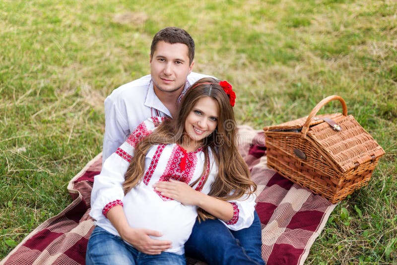 Os pares grávidos novos bonitos vestiram-se no estilo ucraniano nacional que tem o piquenique no parque do outono Maternidade e h imagens de stock Os pares grávidos novos bonitos vestiram-se no estilo ucraniano nacional que tem o piquenique no parque do outono Maternidade e h imagens de stock