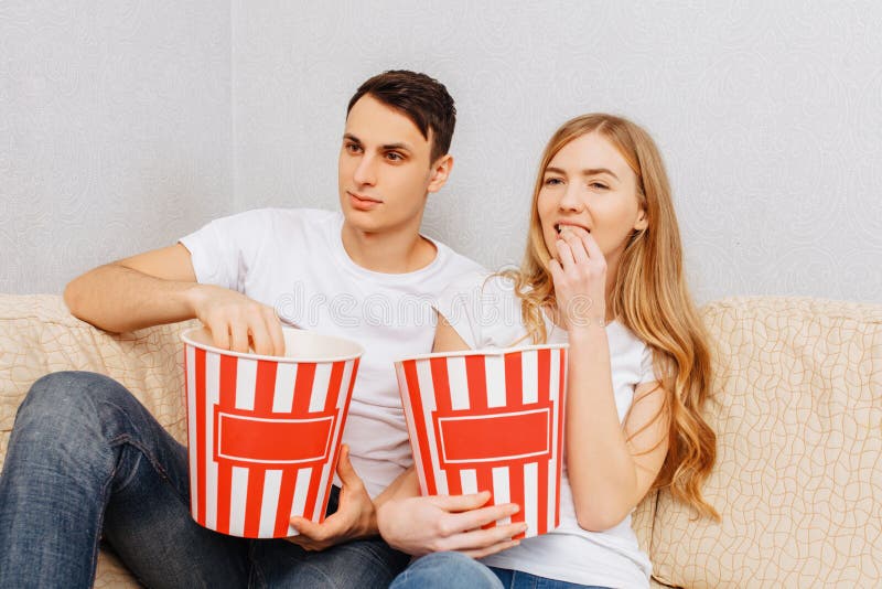 Os pares novos bonitos, homem e mulher, olham filmes e para comer a pipoca, sentando-se em casa no sofá imagens de stock Os pares novos bonitos, homem e mulher, olham filmes e para comer a pipoca, sentando-se em casa no sofá imagens de stock