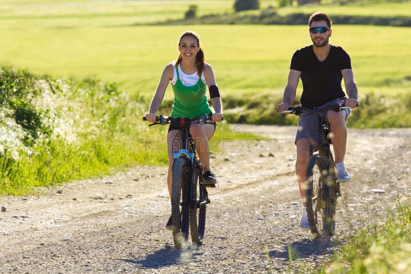 Os pares novos felizes em uma bicicleta montam no campo imagem de stock Os pares novos felizes em uma bicicleta montam no campo imagem de stock