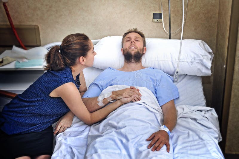 Os pares novos na sala de hospital equipam o encontro na mulher preocupada cama que guarda seu inquietação da mão imagem de stock Os pares novos na sala de hospital equipam o encontro na mulher preocupada cama que guarda seu inquietação da mão imagem de stock