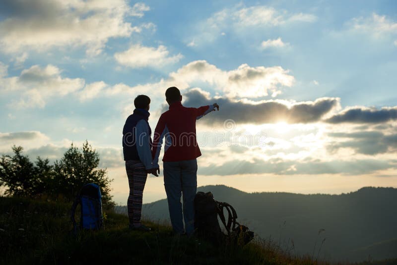 Os turistas da vista traseira dois estão estando sobre a montanha contra o céu nebuloso no por do sol fotografia de stock royalty free Os turistas da vista traseira dois estão estando sobre a montanha contra o céu nebuloso no por do sol fotografia de stock royalty free