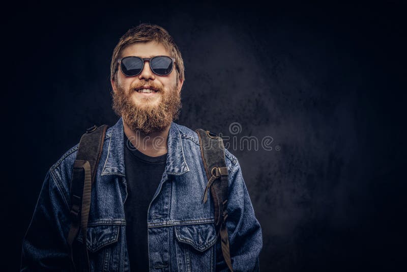 Os óculos de sol vestindo de sorriso e a trouxa do indivíduo farpado do moderno vestiram-se no revestimento das calças de brim em imagem de stock Os óculos de sol vestindo de sorriso e a trouxa do indivíduo farpado do moderno vestiram-se no revestimento das calças de brim em imagem de stock
