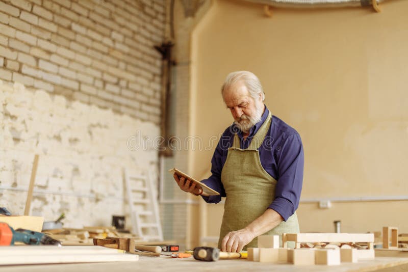 Oude mens met grijze haar, snor en baard die met tablet in de garage werken stock foto's Oude mens met grijze haar, snor en baard die met tablet in de garage werken stock foto's