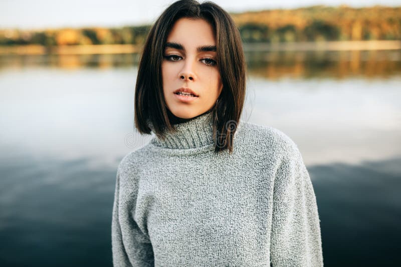 Outdoor close-up portrait of a beautiful young woman wearing grey sweater, posing on nature background. Pretty female looking at