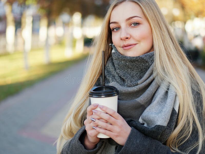 Outdoor close up shot of 20s year stylish blonde woman in scarf on lunch break drinking coffee or tea to go. Autumn urban