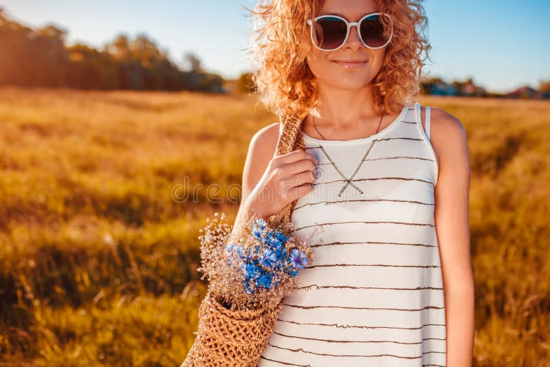 Outdoor fashion portrait of beautiful young woman with red curly hair holding bag with flowers. Summer outfit