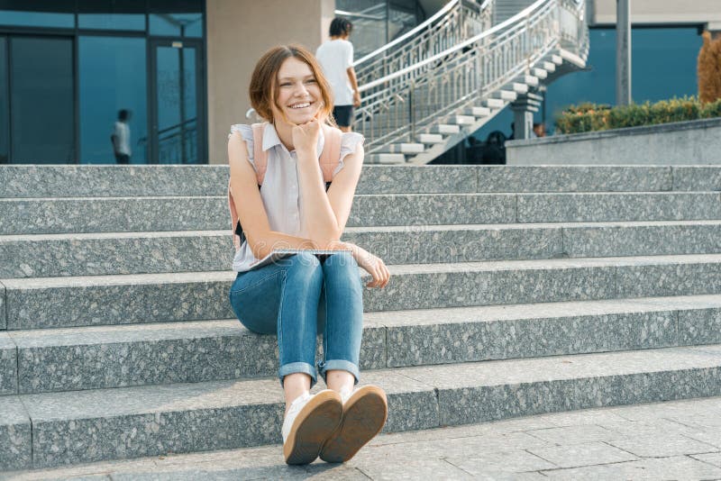 Outdoor portrait of young beautiful smiling student girl with backpack, sitting on the steps in the city