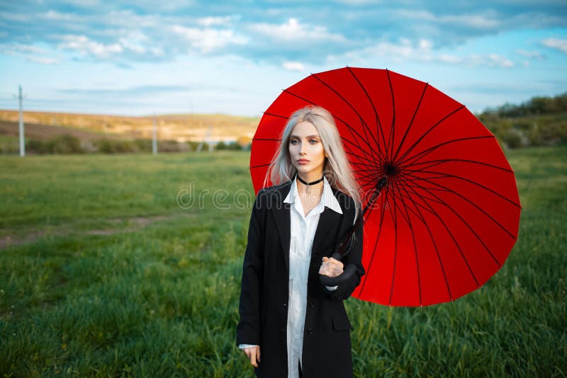 Outdoor portrait of young blonde cute girl, holding red umbrella, wearing black suit. Background of cloudy sky and green field.