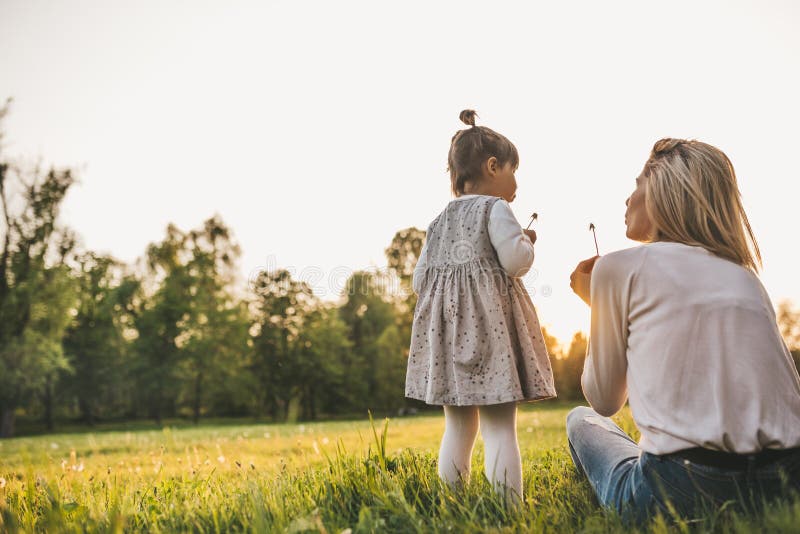 Outdoor rear view of happy girl kid playing with her mother outside. Portrait of woman and her cute child blowing dandelion in the