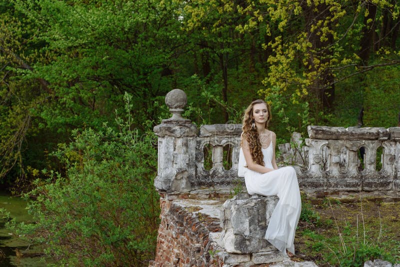 Outdoor summer portrait of young pretty cute girl. Beautiful woman posing at old bridge. in white dess siting near stone railing.