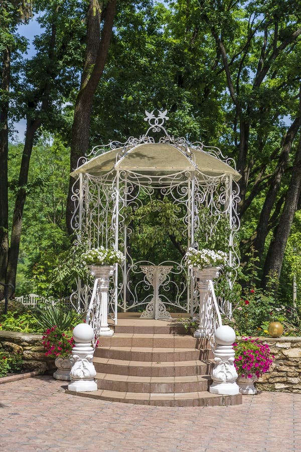 Outdoor white metal gazebo with roof in a garden against green trees