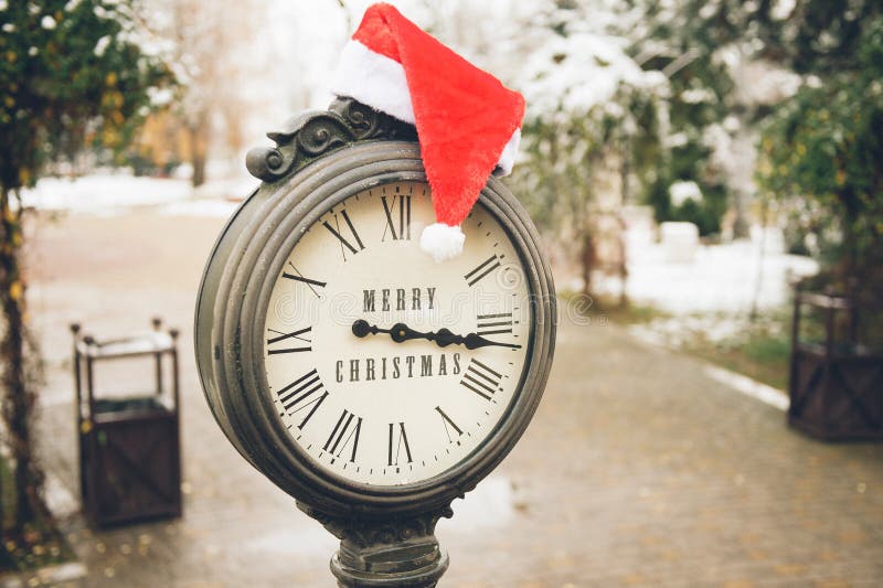 Outdoor winter concept of merry christmas holiday eve old iron clock with title and santa red white hat on it on snow street of