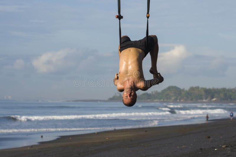 Outdoors portrait of  man practicing aero yoga workout at the beach hanging from rope swing above the sea training body balance
