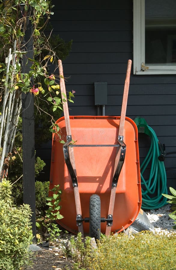 A still life of gardening tools, focusing on an orange wheel barrow leaning against a house, with a coiled green garden hose, and mulch. A still life of gardening tools, focusing on an orange wheel barrow leaning against a house, with a coiled green garden hose, and mulch.
