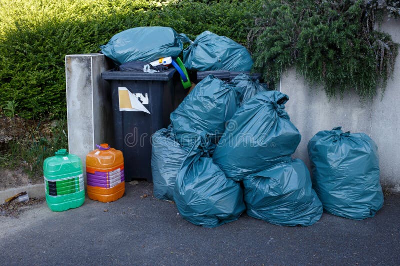 Overflowing outdoor garbage bin with large pile of green trash bags and plastic canisters near concrete wall and stock images
