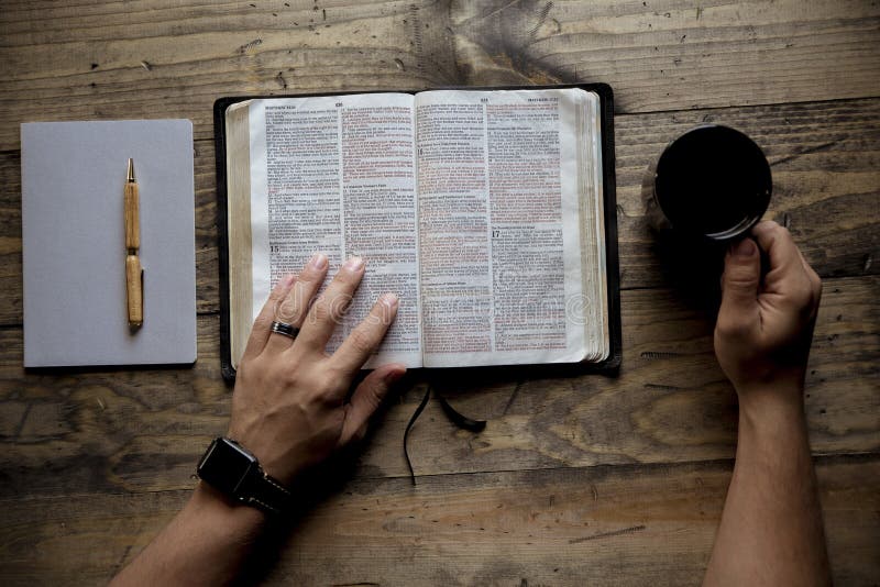 An overhead shot of a male holding his coffee mug and reading a book on a wooden surface