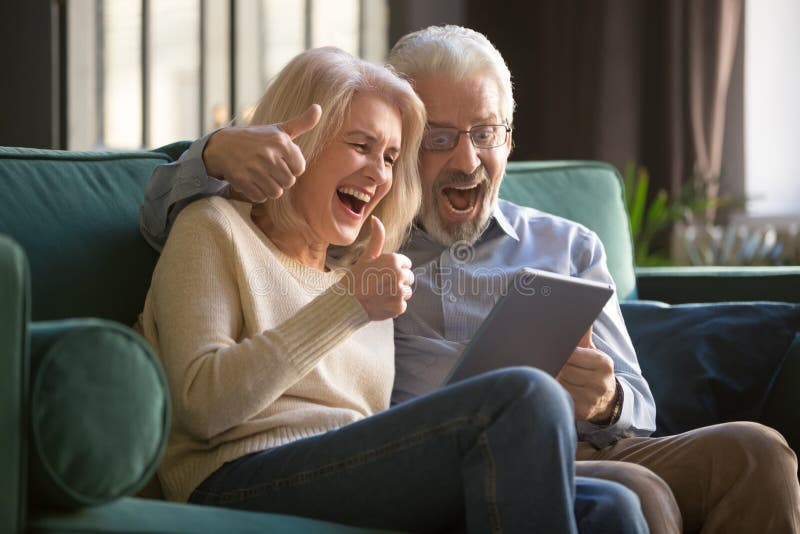 Overjoyed happy senior aged family couple screaming, celebrating online win, success, excited and surprised by perfect news, using tablet, looking at screen, sitting at home on sofa in living room. Overjoyed happy senior aged family couple screaming, celebrating online win, success, excited and surprised by perfect news, using tablet, looking at screen, sitting at home on sofa in living room