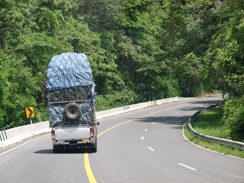 Overloaded mini truck with spare tyre hanging backside on a lonely rural narrow road in tropical lands with green trees and hills.