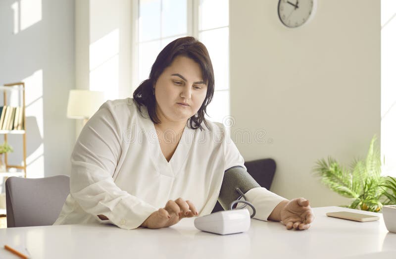 Overweight brunette woman measuring pressure while sitting at table at home