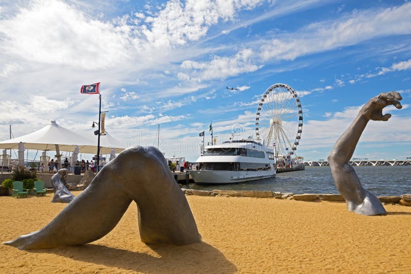 OXON HILL, MARYLAND, USA - SEPTEMBER 11, 2016: Awakening, a sculpture at National Harbor in front of pier and Ferris wheel. A famous statue of a giant embedded in the earth created by J. Seward Johnson Jr. OXON HILL, MARYLAND, USA - SEPTEMBER 11, 2016: Awakening, a sculpture at National Harbor in front of pier and Ferris wheel. A famous statue of a giant embedded in the earth created by J. Seward Johnson Jr