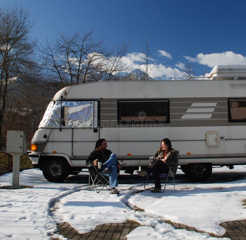 A couple sit outside their motorhome with hot drinks on a cold snowy morning in the mountains. They are looking at each other and laughing. A couple sit outside their motorhome with hot drinks on a cold snowy morning in the mountains. They are looking at each other and laughing.