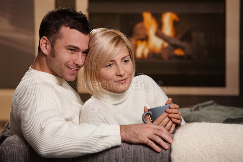 Young love couple hugging on sofa in front of fireplace at home, looking away, smiling. Young love couple hugging on sofa in front of fireplace at home, looking away, smiling.