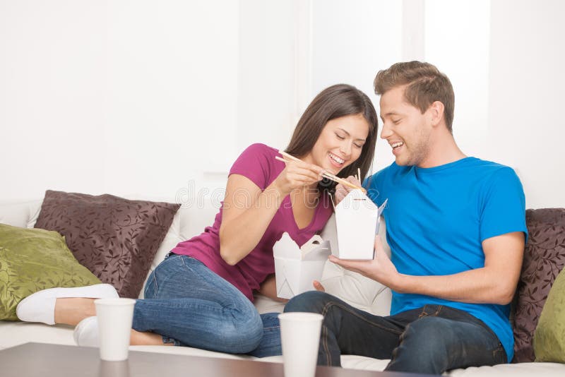 Beautiful couple eating Asian food from food containers while sitting close to each other on the couch. Beautiful couple eating Asian food from food containers while sitting close to each other on the couch