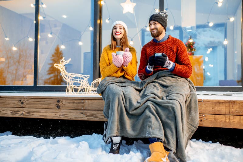Young couple warming up with plaid and hot drinks sitting on the terrace of the modern house in the mountains durnig the winter holidays. Young couple warming up with plaid and hot drinks sitting on the terrace of the modern house in the mountains durnig the winter holidays