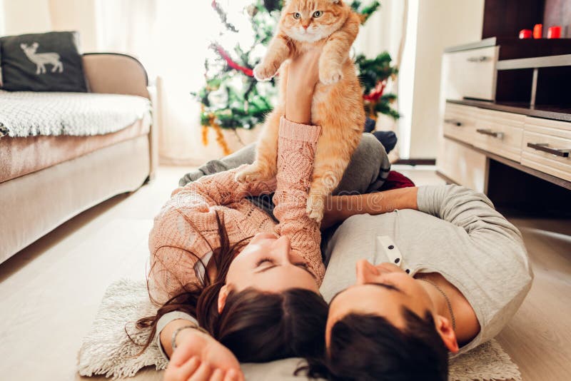 Paar in liefde die door Kerstboom liggen en met kat thuis spelen Man en vrouwen het opheffen huisdier in handen stock foto's Paar in liefde die door Kerstboom liggen en met kat thuis spelen Man en vrouwen het opheffen huisdier in handen stock foto's