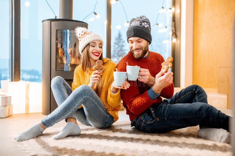 Young couple dressed in bright sweaters sitting together with hot drinks near the fireplace in the modern house in the mountains durnig winter time. Young couple dressed in bright sweaters sitting together with hot drinks near the fireplace in the modern house in the mountains durnig winter time