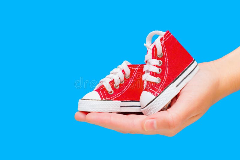 Pair of miniature red high-top canvas shoes with white laces and rubber soles held in the palm of a hand. E-commerce,online shopping,fashion-related concepts targeting kids' footwear. Pair of miniature red high-top canvas shoes with white laces and rubber soles held in the palm of a hand. E-commerce,online shopping,fashion-related concepts targeting kids' footwear