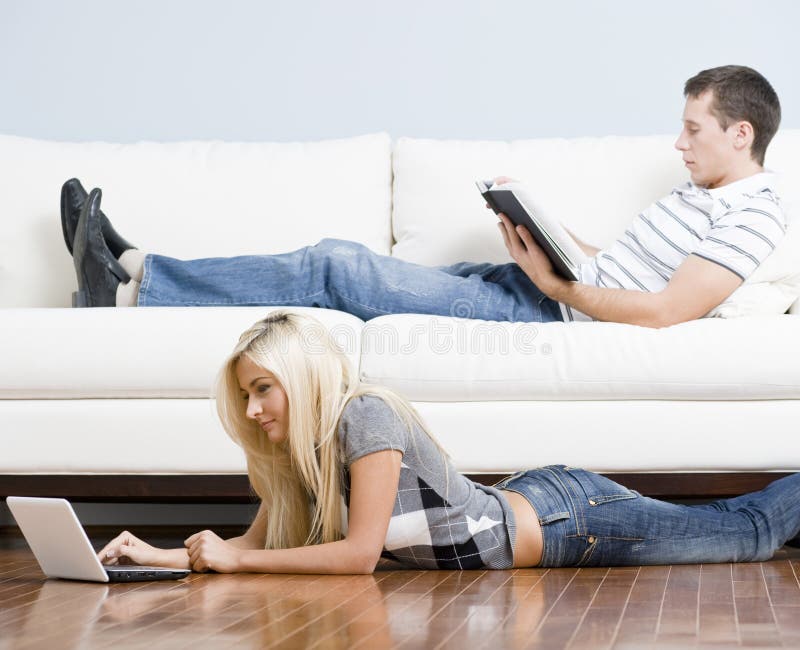 Man reads on a couch while woman stretches out on the floor with her laptop. Horizontal format. Man reads on a couch while woman stretches out on the floor with her laptop. Horizontal format.