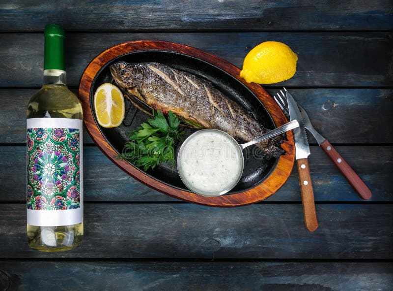 Padella con il pesce saporito della trota su fondo di legno con una bottiglia dei verdi, della salsa e del limone del vino fotografia stock libera da diritti Padella con il pesce saporito della trota su fondo di legno con una bottiglia dei verdi, della salsa e del limone del vino fotografia stock libera da diritti