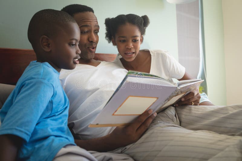 Padre afroamericano con i suoi bambini che leggono un libro di fiabe sul letto in camera da letto