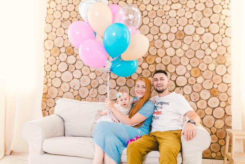 Padre de familia, mujer joven y bebé sentándose en el sofá dentro con la pared decorativa de madera y los globos multicolores con fotografía de archivo libre de regalías Padre de familia, mujer joven y bebé sentándose en el sofá dentro con la pared decorativa de madera y los globos multicolores con fotografía de archivo libre de regalías
