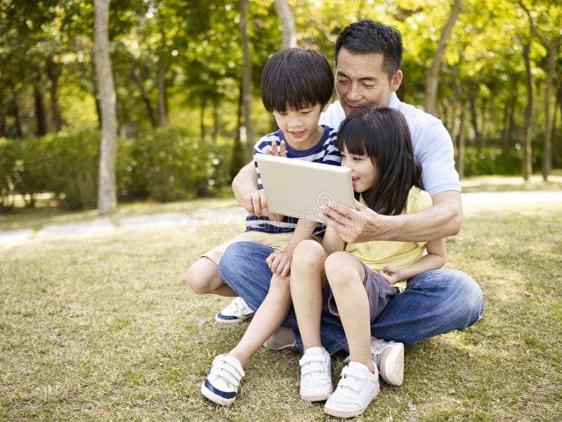 Asian father and two children sitting on grass looking at tablet computer, outdoor in a park. Asian father and two children sitting on grass looking at tablet computer, outdoor in a park.