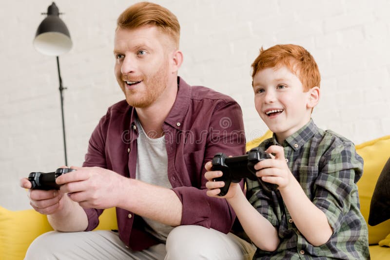 Padre e hijo sonrientes que se sientan en el sofá y que juegan con las palancas de mando fotografía de archivo